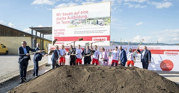 Eine Gruppe von Personen hebt mit Schaufeln Erde an einer Baustelle. Große Werbetafel mit Text: Wir bauen auf eine starke Ausbildung. Im Team. karriere.strabag.com STRABAG TEAMS WORK. Jetzt bewerben.