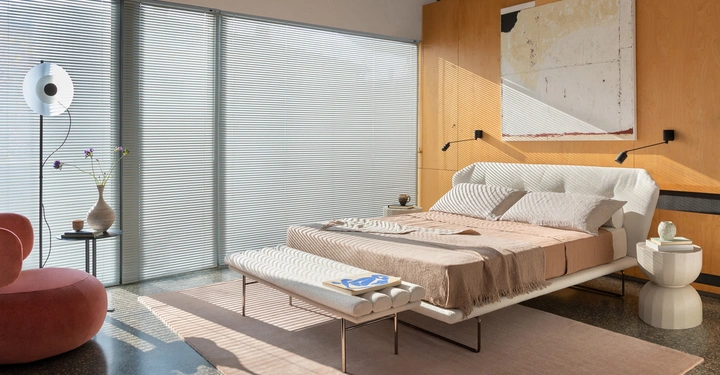 Ein großes Bett liegt in einem modernen Schlafzimmer mit Holzwand-Akzent vertikale Jalousien am Fenster einem runden Stuhl in Rosa einem Beistelltisch einer Vase und einer Stehlampe.