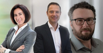 Drei Personen lächeln in die Kamera Frau in grauem Blazer verschränkt die Arme Mann in dunklem Anzug steht aufrecht bärtiger Mann mit Brille blickt in frischem Raum mit hellgrauem Hintergrund.