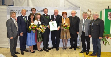 Gruppe von zehn Menschen steht in einer Reihe, zwei halten Blumensträuße und ein Mann zeigt eine Urkunde. Im Hintergrund ein Banner mit dem Text EINFACH RICHTIG BAUEN.