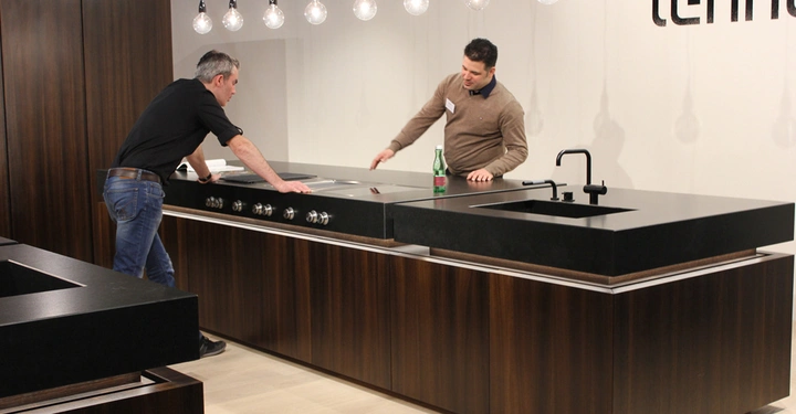 Galerie-Bild 19: Zwei Männer unterhalten sich und gestikulieren an einer schwarzen Kücheninsel in einem modernen Ausstellungsraum mit hölzernen Schränken und hängenden Glühbirnen im Hintergrund. Text: lehner