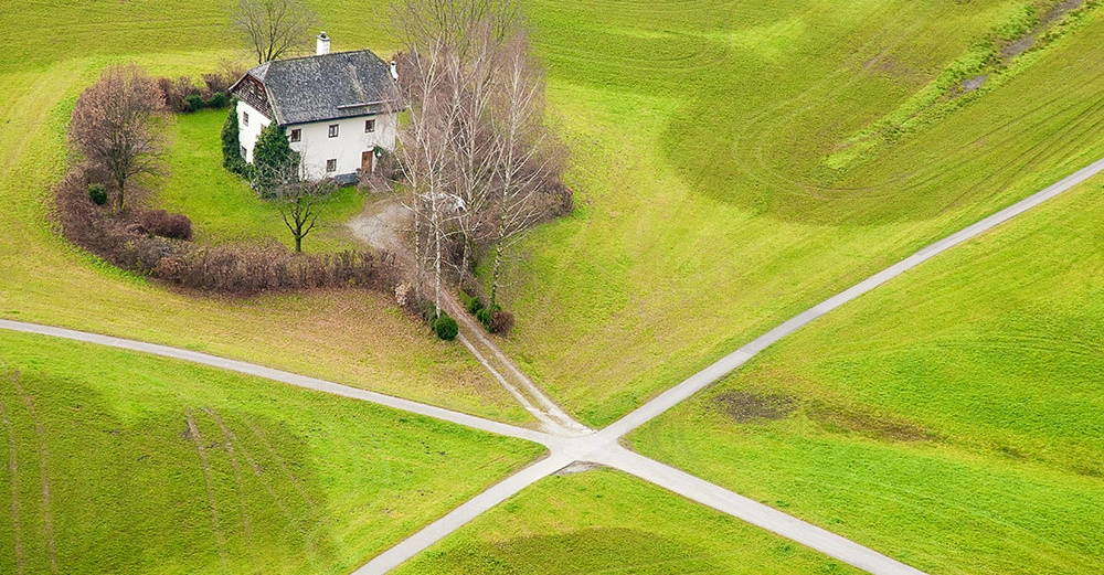 Ein Haus steht in einem kleinen Bereich mit Bäumen und Büschen im Zentrum eines grünen Feldes mit sich kreuzenden Wegen