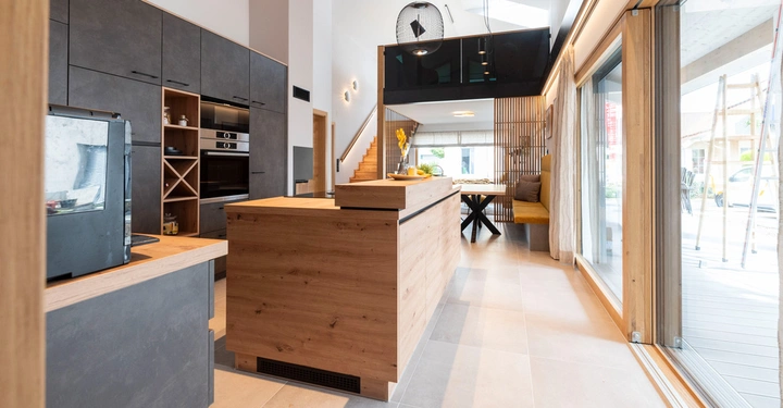 Holzinsel in moderner Küche mit integriertem Herd und Ofen während große Fenster das Tageslicht in den offenen Wohn-Essbereich einlassen. Treppe im Hintergrund führt zu einem Zwischengeschoss