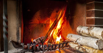 Ein brennendes Feuer im Kamin lodert und erhellt den Raum mit Holzscheiten und Metallgitter, umgeben von Ziegelwänden und einem gemauerten Kamin.