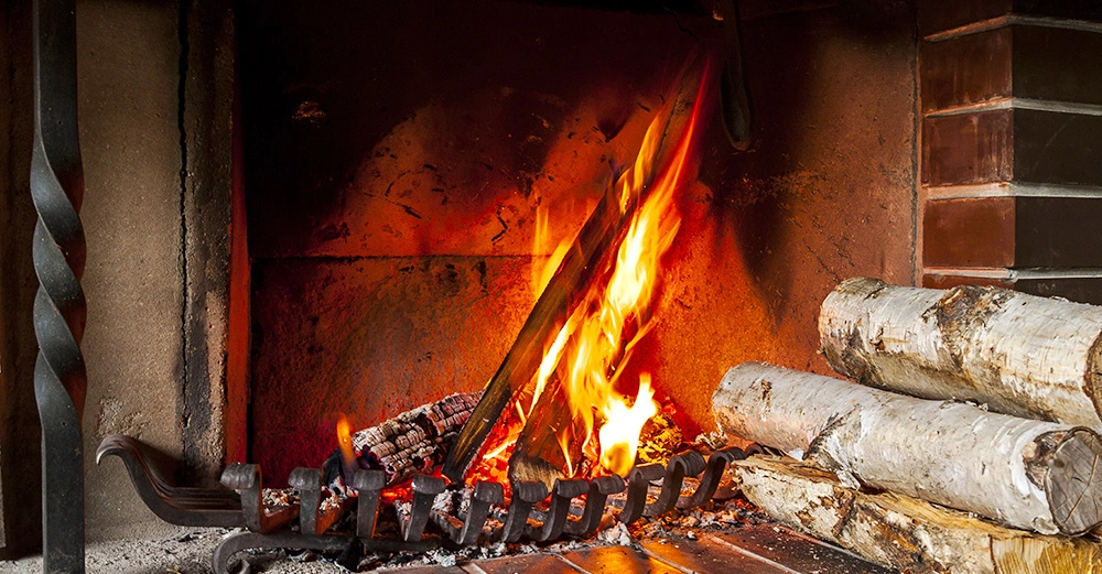 Ein brennendes Feuer im Kamin lodert und erhellt den Raum mit Holzscheiten und Metallgitter, umgeben von Ziegelwänden und einem gemauerten Kamin.