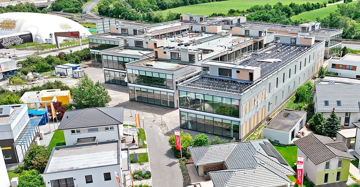Luftaufnahme des neuen Bauzentrums in der Blauen Lagune. 5 quaderförmige Gebäude mit viel Stahl und Glas inmitten der Fertighäuser. Grüne Wiese im Hintergrund. 