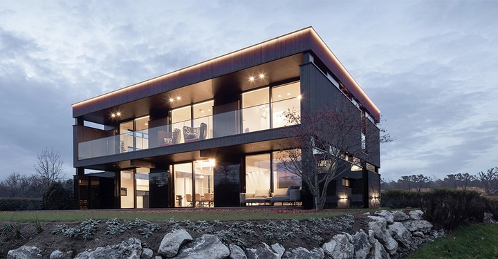 Galerie-Bild 2: Abendliche Stimmung mit innen beleuchtetem Haus mit 6 großen Fenstern udn Falchdach. Zu sehen ist das Haus, eine Wiese mit kleiner Mauer im Vordergrund und wolkigem Himmel im Hintergrund. 