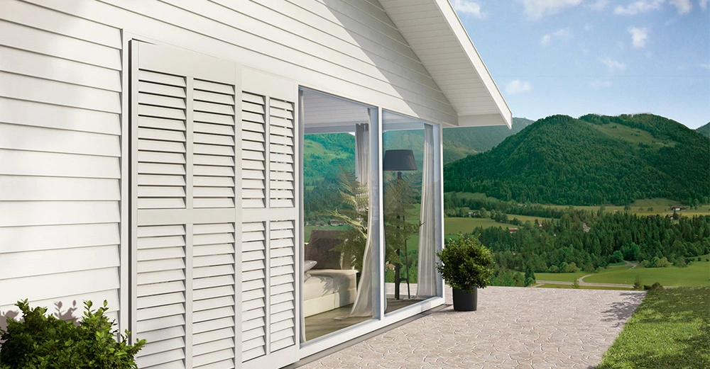 Ein weißes Haus mit großen Fenstern steht auf einer Terrasse im Freien umgeben von grünen Hügeln und blauem Himmel. Ein Blumentopf steht auf der Terrasse.