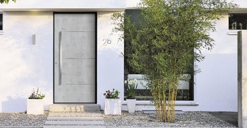 Graue Haustür steht auf grauem Steinweg vor weißem Gebäude Nummer 2b Vor der Tür und Fenstern sind Blumen und Sträucher gepflanzt.