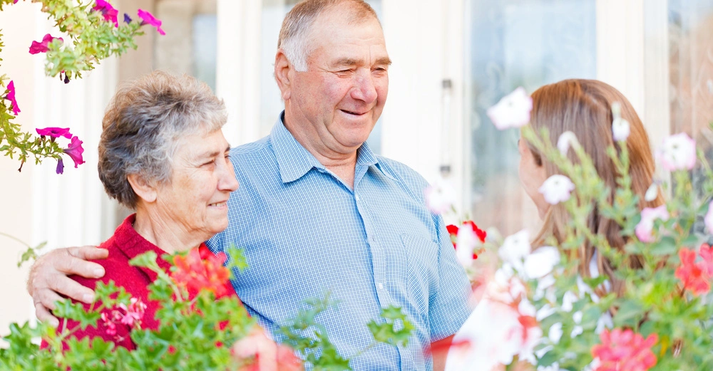 Ein älteres Paar umarmt sich und unterhält sich mit einer jüngeren Frau im Garten umgeben von bunten Blumen vor einem Haus mit Fenstern.