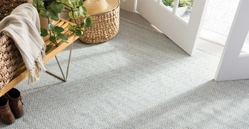 Pflanzen und ein gefalteter Schal liegen auf einer Holzbank auf einem gemusterten Teppich in einem hellen Eingangsbereich mit offenen Türen und einem Weidenkorb.