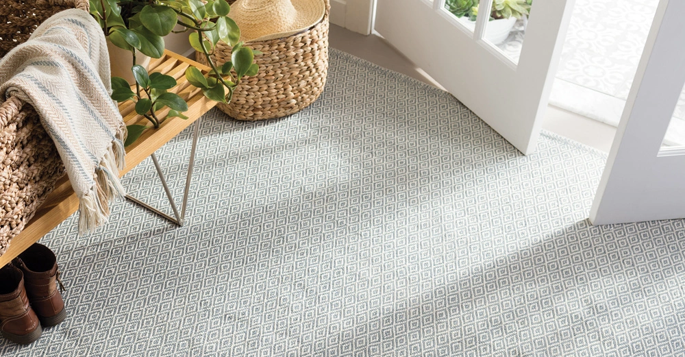 Pflanzen und ein gefalteter Schal liegen auf einer Holzbank auf einem gemusterten Teppich in einem hellen Eingangsbereich mit offenen Türen und einem Weidenkorb.