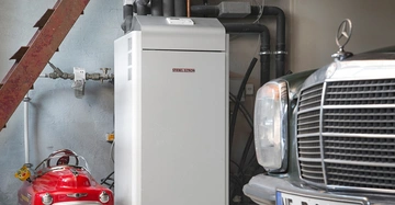 Eine Wärmepumpe steht still in einer Garage neben einem Spielzeugauto und einem klassischen Auto mit dem Kennzeichen NE D1415H. Beschriftung auf der Wärmepumpe lautet STIEBEL ELTRON.