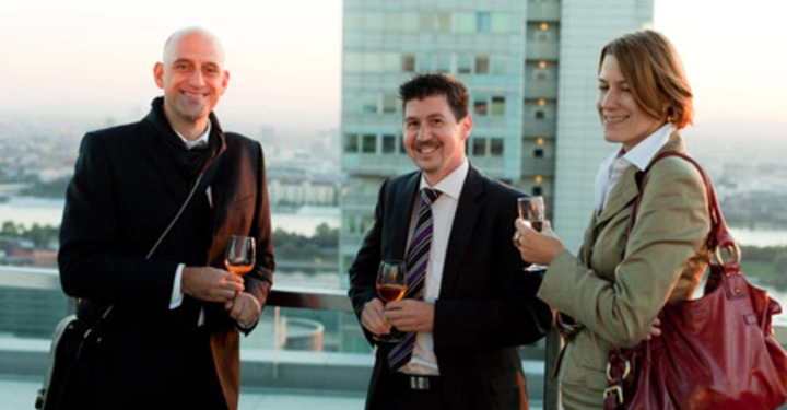 Galerie-Bild 25: Drei Personen halten Weingläser und lächeln auf einer Dachterrasse mit Stadtblick Hinter ihnen steht ein modernes Hochhaus In der Ferne ist ein Fluss und eine Stadtlandschaft zu sehen