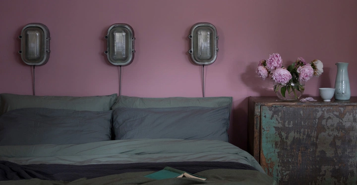Ein Bett mit grauer Bettwäsche steht vor einer rosa Wand mit drei Wandlampen, daneben eine Kommode mit einer Vase voller rosa Blumen und einer weißen Tasse in einem Schlafzimmer.