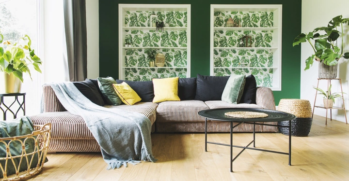 Ein beigefarbener Sofa steht vor einer grünen Wand mit Regalnischen voller Pflanzenmustertapeten im Wohnzimmer ein schwarzer Couchtisch steht vor dem Sofa links und rechts sind Zimmerpflanzen.