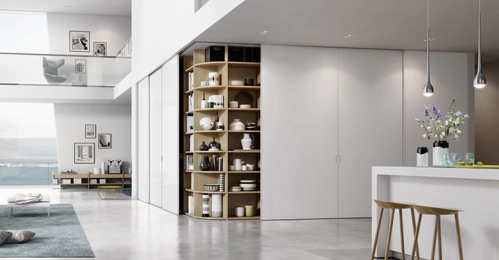 Galerie-Bild 4: Regale zeigen Geschirr in einem modernen Wohnzimmer mit einer offenen Küche Dekoration bestehend aus Blumen auf einer weißen Kücheninsel mit Barhockern große Fenster bieten Blick auf das Meer und Berge