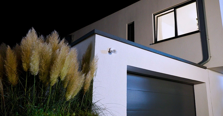 Perspektivische Ansicht eines Hauses mit vorgebauter Garage, im oberen Stockwerk brennt Licht, im Vordergrund eine Kamera bzw. Bewegungsmelder an der Außenwand