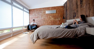 Zwei Personen entspannen in einem modernen Schlafzimmer. Eine Person sitzt in einem Sessel und liest ein Buch, während die andere Person auf einem Hochbett sitzt und eine Tasse hält.