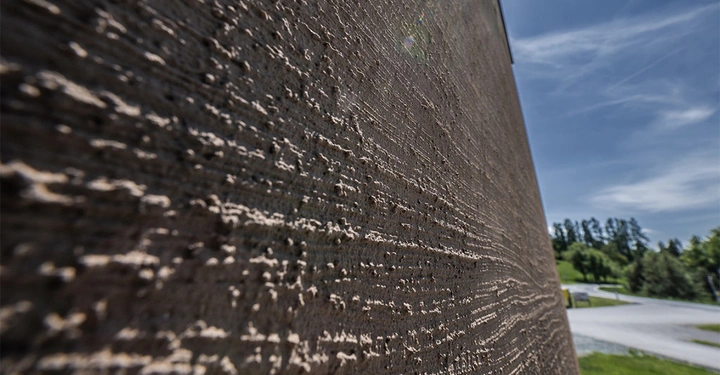Galerie-Bild 3: Braungraue Fassade mit Struktur perpektivisch fotografiert, im Hintergrund blauer Himmel und Wald