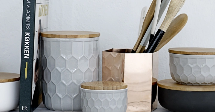 Galerie-Bild 3: Kochutensilien im Kupferbehälter neben grauen Keramikbehältern mit Holzdeckeln auf einer weißen Küchenarbeitsplatte Kochbuch "Timm Vladimirs Køkken" steht links aufrecht.