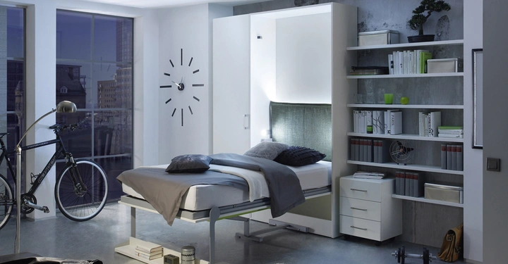 Ein Bett steht in einer modernen Wohnung mit Glasfenstern im Hintergrund, daneben ein Regal mit Büchern und Dekorationen. Links steht ein Fahrrad.