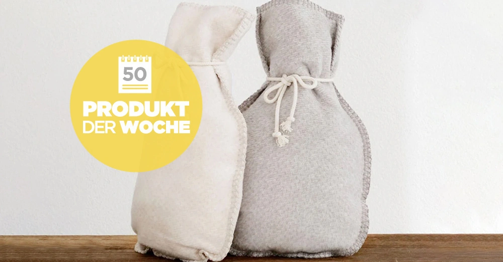 Zwei Wärmflaschen stehen auf einem Holztisch vor einer weißen Wand. Ein gelber Kreis mit Text überlagert das Bild. Text im Kreis: 50 PRODUKT DER WOCHE.