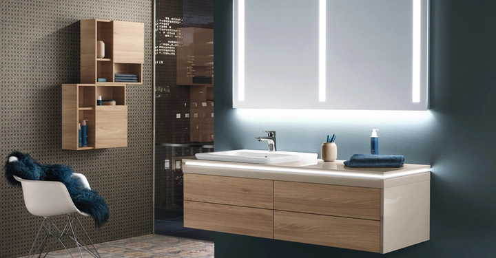 Ein Badezimmerspiegel mit LED-Beleuchtung hängt über einem Waschbecken auf einem Holzschrank; im Hintergrund stehen ein Stuhl mit Pelzdecke und hölzerne Regale an einer gemusterten Wand.