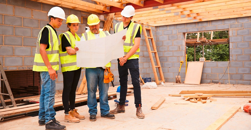 Bauarbeiter tragen Schutzhelme und Sicherheitswesten und betrachten in einer unfertigen Baustelle eine Bauzeichnung. Im Hintergrund befinden sich Wände aus Ziegelsteinen, Holz und Bauwerkzeuge.