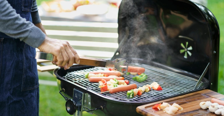 Eine Hand hält eine Grillzange und dreht Würstchen auf einem dampfenden Grill, umgeben von Paprika und anderen Gemüsesorten im Hintergrund eine Gartenlandschaft.