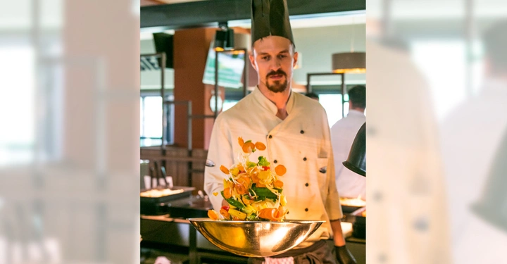 Galerie-Bild 7: Ein Koch trägt eine weiße Kochjacke und einen schwarzen Hut hält eine Schüssel mit geschnittenem Gemüse in einem Restaurant mit moderner Innenausstattung im Hintergrund.
