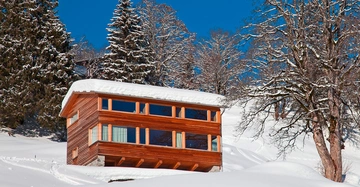 Ein verschneites Holzhaus steht ruhig auf einem Hügel umgeben von schneebedeckten Bäumen und klarem blauem Himmel