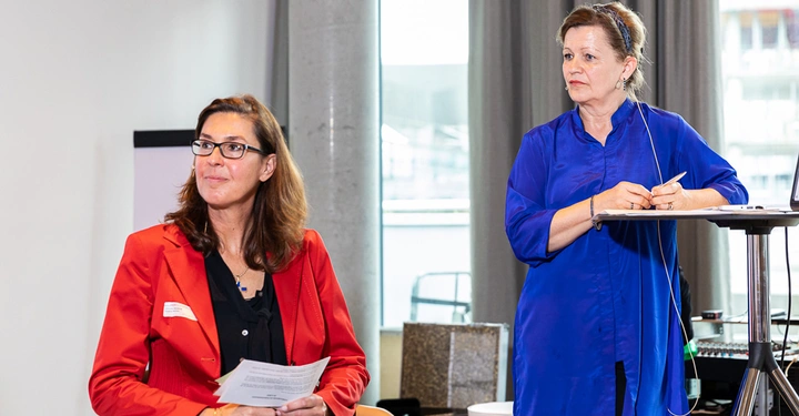 Galerie-Bild 5: Zwei Frauen sitzen und stehen vor einem Tisch in einem Besprechungsraum Eine Frau in rotem Blazer hält Papiere Die andere Frau in blauem Hemd steht am Tisch und hält einen Stift