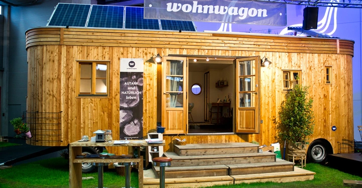Galerie-Bild 1: Wohnwagen mit Holzverkleidung steht in einer Ausstellungshalle Solarpanels oben Banner mit dem Text wohnwagon Tafel mit Text AUTARK UND NATÜRLICH LEBEN kleiner Tisch mit Stühlen und Dekor.