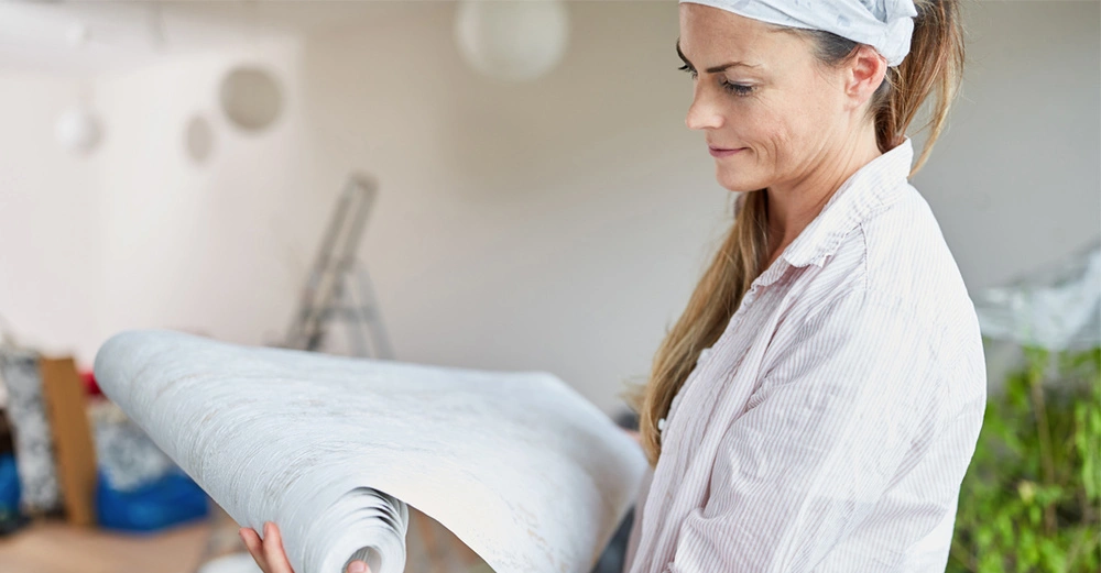 Eine Frau hält eine Rolle Tapete. Sie steht in einem Raum, der renoviert wird, mit Leitern und abgedeckten Möbeln im Hintergrund.