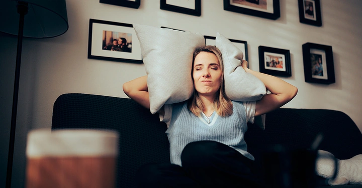 Eine Frau hält sich aufgrund zu hoher Lautstärke der Nachbarn über ihr mit zwei Kissen die Ohren zu.