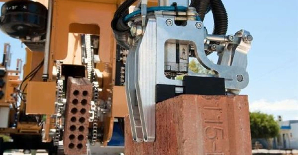 Ein mechanischer Greifer hebt einen Ziegelstein an einem sonnigen Tag auf einer Baustelle hoch. Im Hintergrund sind Maschinen und Bäume zu sehen. Text auf dem Ziegelstein 115 E3.