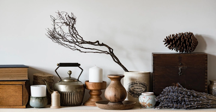 Verschiedene dekorative Gegenstände wie Kerzen Kannen Vasen Zweige Bücher und ein Kiefernzapfen auf einem Regal in einem minimalistischen Raum