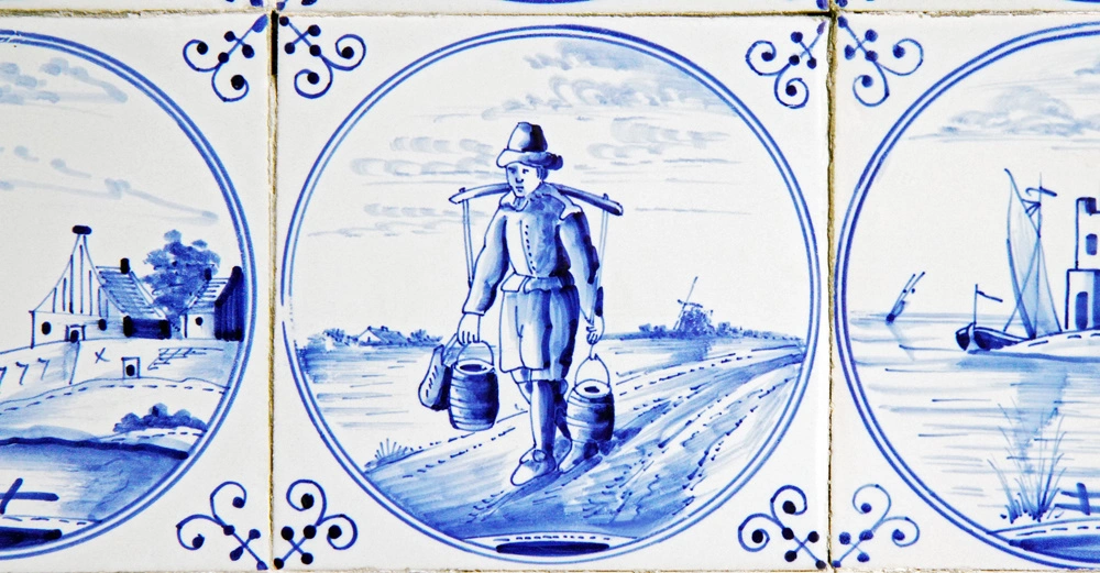 Kachel mit einem Mann der zwei Eimer trägt ländliche Umgebung mit Häusern und Segelbooten verziert mit blauen Ornamenten und dargestellten Szenerien im Kreisrahmen auf weißem Hintergrund.