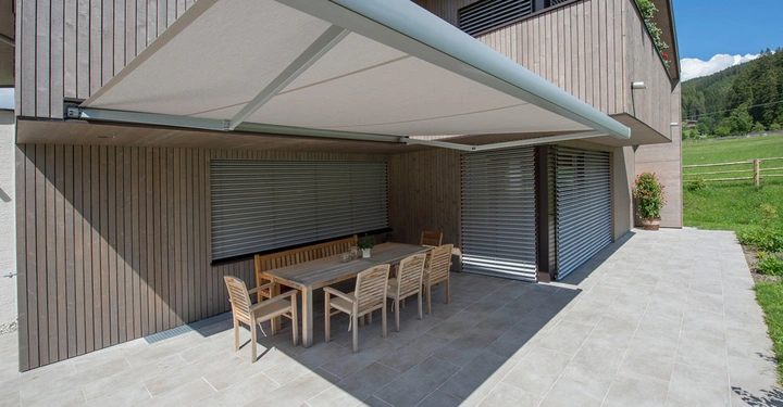 Holztisch mit sechs Stühlen steht auf einer gefliesten Terrasse unter einer großen Markise vor einem modernen Holzhaus mit geschlossenen Jalousien; umgeben von Wiese und Bäumen