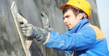 Bauarbeiter mit gelbem Helm verputzt Außenfassade mit Kelle, grauer Unterputz wird aufgebracht