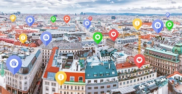 Mehrere bunte Standortmarkierungen schweben über Gebäude in einer städtischen Umgebung. Die Markierungen zeigen Symbole wie Häuser, Herzen, Sterne und Ausrufezeichen. Die Stadtlandschaft erstreckt sich unter einem bewölkten Himmel.