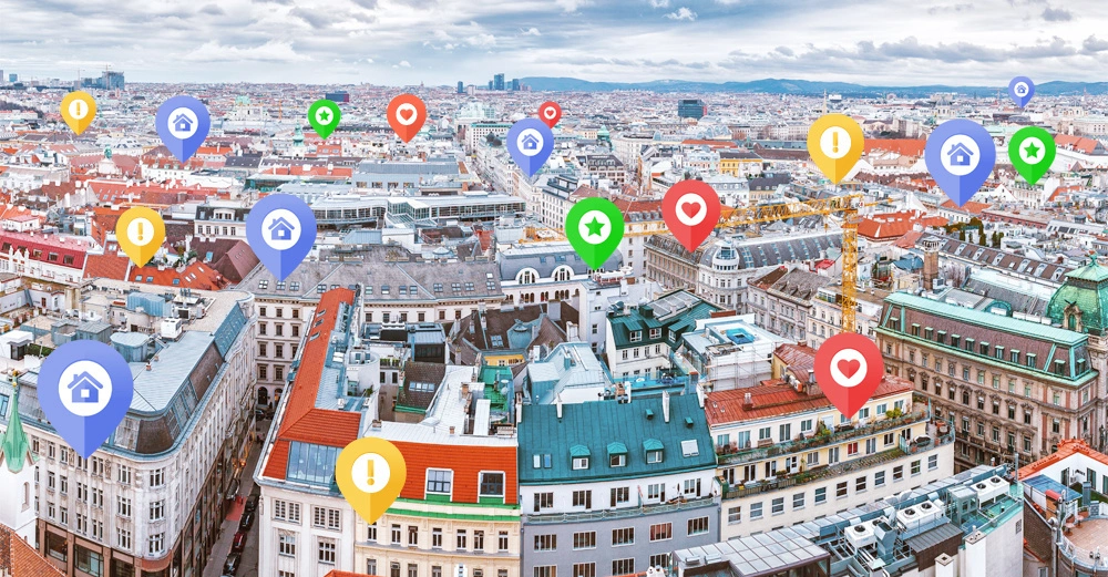 Mehrere bunte Standortmarkierungen schweben über Gebäude in einer städtischen Umgebung. Die Markierungen zeigen Symbole wie Häuser, Herzen, Sterne und Ausrufezeichen. Die Stadtlandschaft erstreckt sich unter einem bewölkten Himmel.