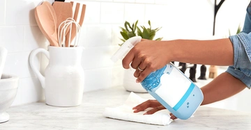 Hand sprüht eine Flüssigkeit aus einer Sprühflasche auf eine Arbeitsfläche in einer hellen Küche mit Küchenutensilien und Pflanzen im Hintergrund