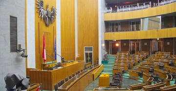 Holzgetäfelte Rednertribüne mit österreichischer und EU-Flagge und großem Metalladler an der Wand vor geschwungenem Raum mit leeren Holzbänken und Stuhlreihen in einem Parlamentssaal.
