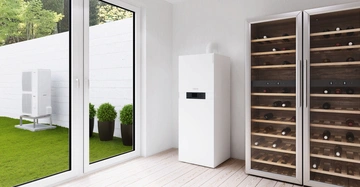 Weißer Heizungskessel steht neben zwei Weinkühlschränken in einem hellen Raum mit Holzboden große Fenster zeigen einen grünen Garten mit Pflanzen und Klimagerät.