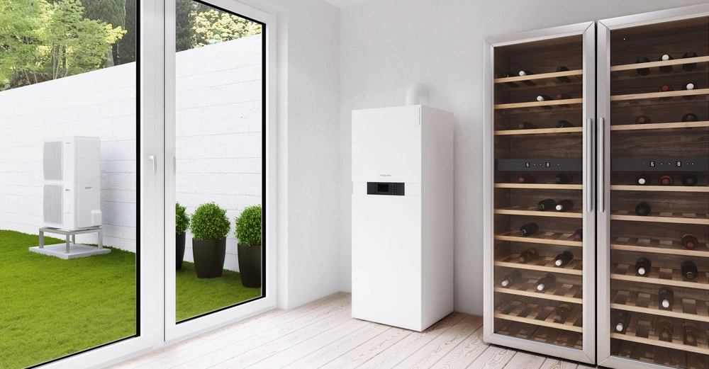 Weißer Heizungskessel steht neben zwei Weinkühlschränken in einem hellen Raum mit Holzboden große Fenster zeigen einen grünen Garten mit Pflanzen und Klimagerät.
