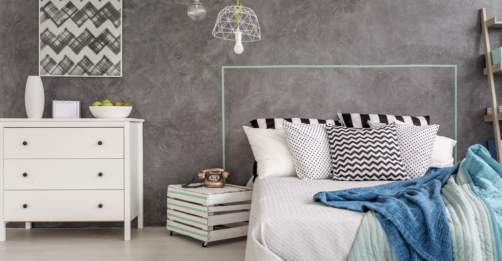 Bett mit gemusterten Kissen und Decken steht vor einer grauen Wand. Links eine weiße Kommode mit dekorativem Zubehör. Hängelampen und abstrakte Kunst schmücken den Raum.