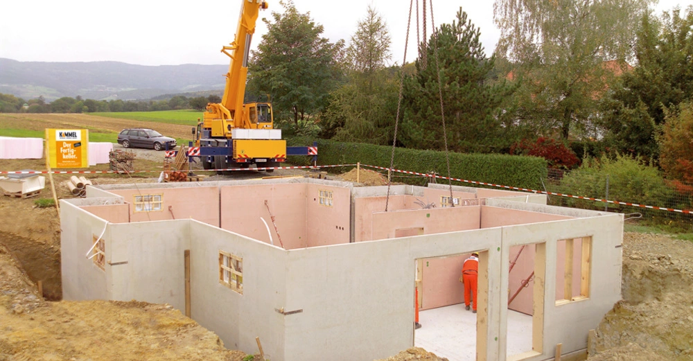 Ein Bauarbeiter richtet Betonteile im Rohbau eines Hauses aus ein Kran hebt Teile in eine Grube im Vordergrund Felder und ein Auto im Hintergrund Wald und Berge Text: KAMMEL Der Fertigkeller