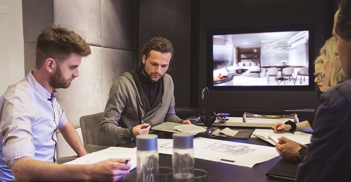Personen arbeiten an einem Tisch mit Architekturplänen in einem Konferenzraum. Ein großer Bildschirm zeigt ein modernes Innenraumdesign. Wasserflaschen stehen auf dem Tisch.